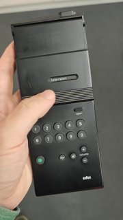 Рука держит универсальный пульт Braun RC1 (1979), вид в ладони: пластик, клавиши 0–9, кнопка 'open' и зелёный индикатор; масштаб и эргономика