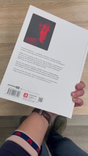 Рука держит книгу в магазине: задняя обложка с красным портретом, логотип издательства, штрихкод и QR‑код на белом фоне, видна часть руки и браслет.