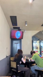 Фото в кафе: настенный вентилятор над дверью с подсветкой, посетители за столом и холодильник с напитками — пример сомнительного дизайна.