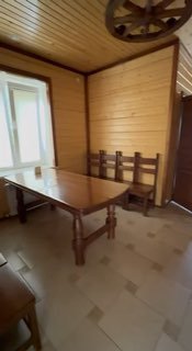 Интерьер предбанника: деревянный стол и несколько стульев в уютной комнате отдыха у окна, светлый деревянный потолок и плиточный пол.