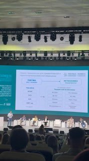 Панель спикеров на сцене форума PreventMed: несколько выступающих на фоне слайда с таблицей и диаграммой, зрители в креслах зала слушают дискуссию.