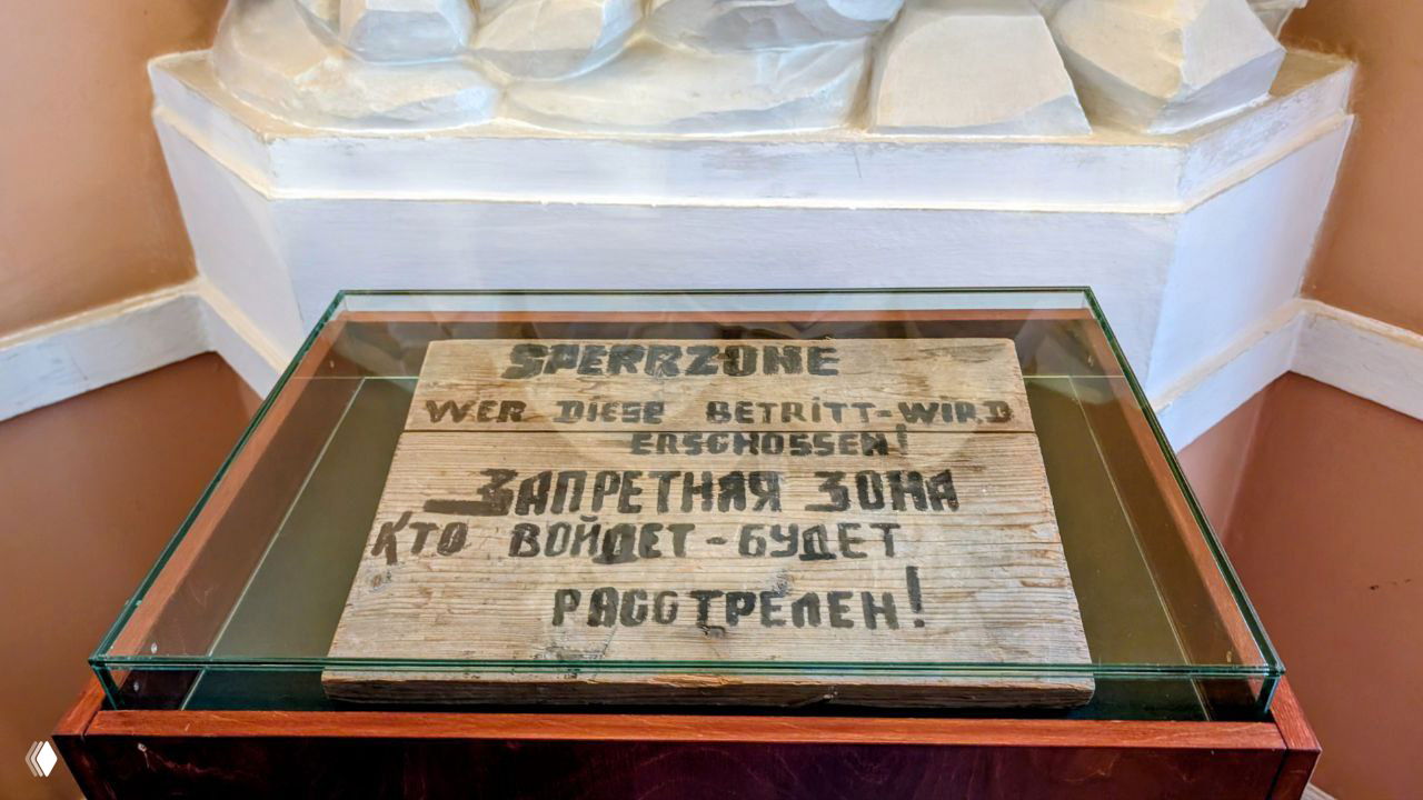 Историческая табличка на немецком и русском с предупреждением об угрозе расстрела, выставленная в витрине как экспонат времен оккупации.