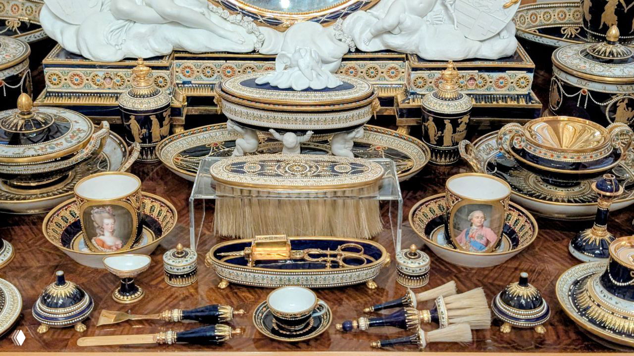 Крупный план фарфоровых предметов на витринном столе: чашки, подносы и аксессуары с портретами и золотыми украшениями.