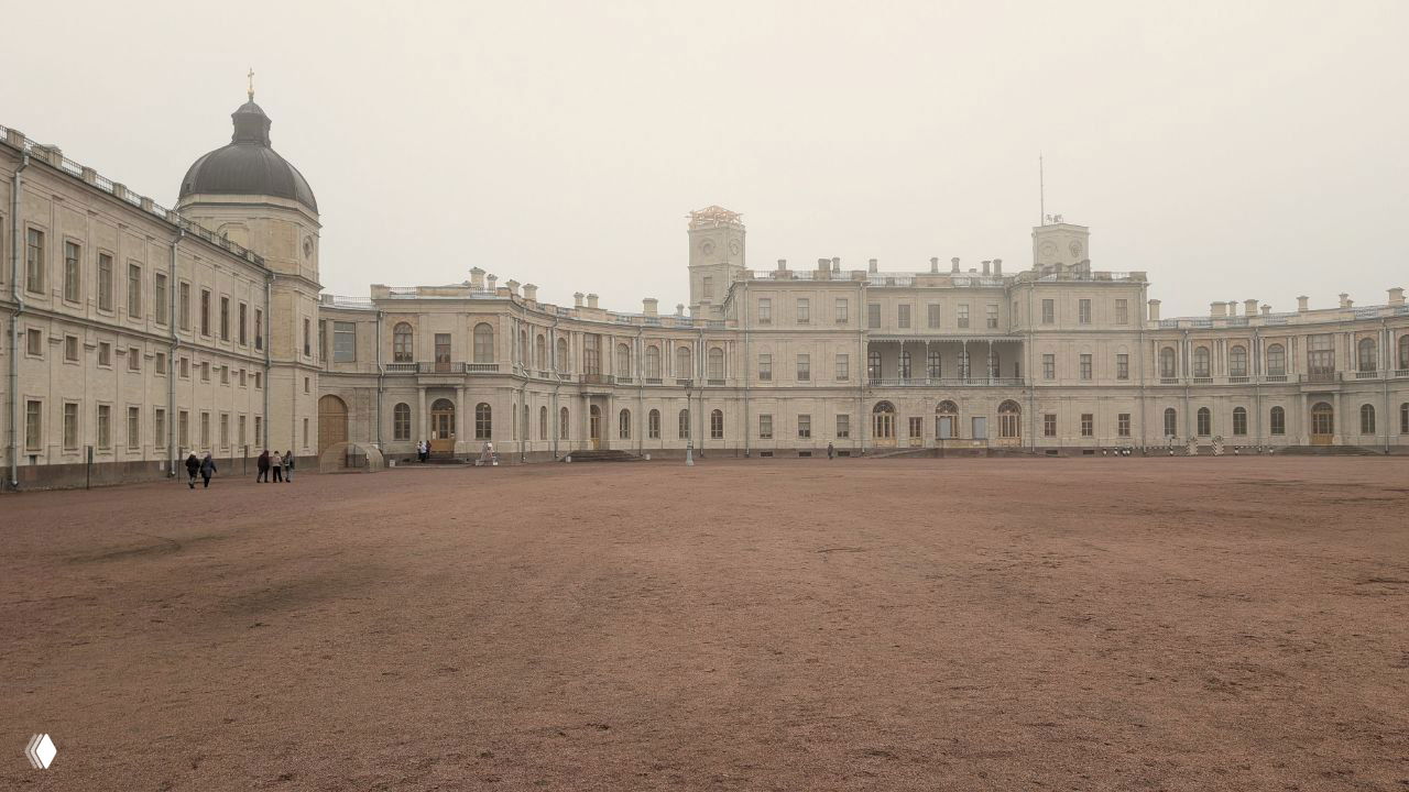 Широкий план дворцово-паркового фасада Гатчины в пасмурный день: дуга зданий, пустая гравийная площадь и туманная перспектива без туристического шума.