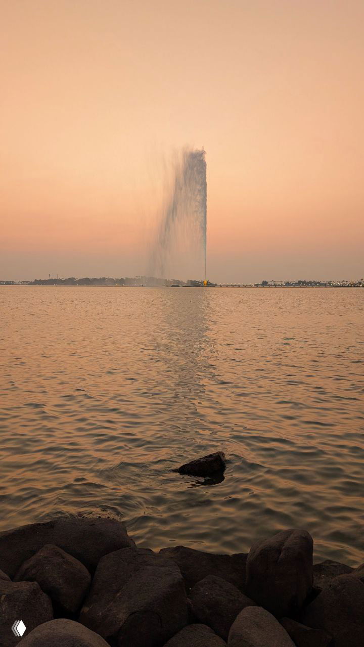 Горизонтальная перспектива на фонтан короля Фахда: высокая струя посреди моря, передний план с камнями и отражением воды при вечернем свете.