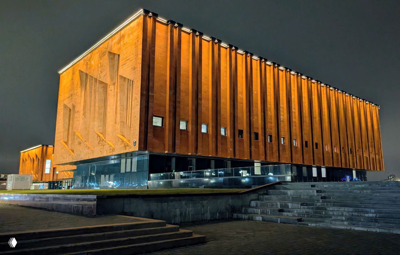 Угловой фасад современной кирпично-коричневой архитектуры библиотеки ночью; крупные рельефные панели и подсветка по контуру крыши.