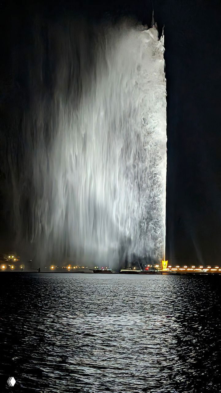 Ночное фотографии фонтанного столба короля Фахда: ярко освещённая мощная струя воды и огни променада на фоне тёмного неба и моря.