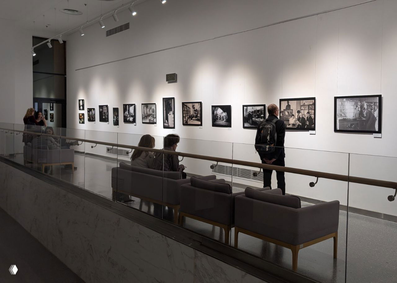 Зал выставки с рядом рамочных фото на стене и посетителями, просматривающими экспозицию в более спокойной части пространства.