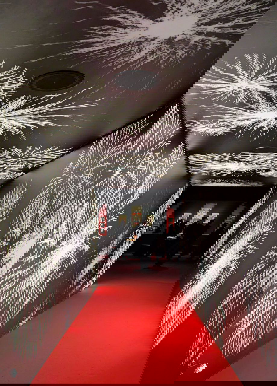 Проход-туннель экспозиции с проекциями белых фейерверков на чёрных стенах и красной дорожкой — световая инсталляция, создающая атмосферу праздника.