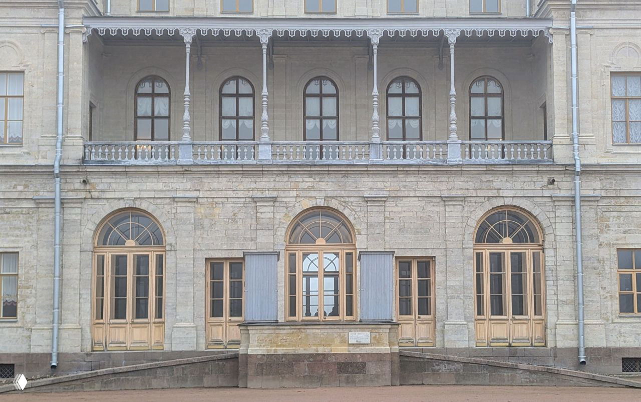 Фрагмент фасада с галереей и арочными окнами Большого Гатчинского дворца: балюстрады, деревянные двери-окна и светлая каменная кладка вблизи.