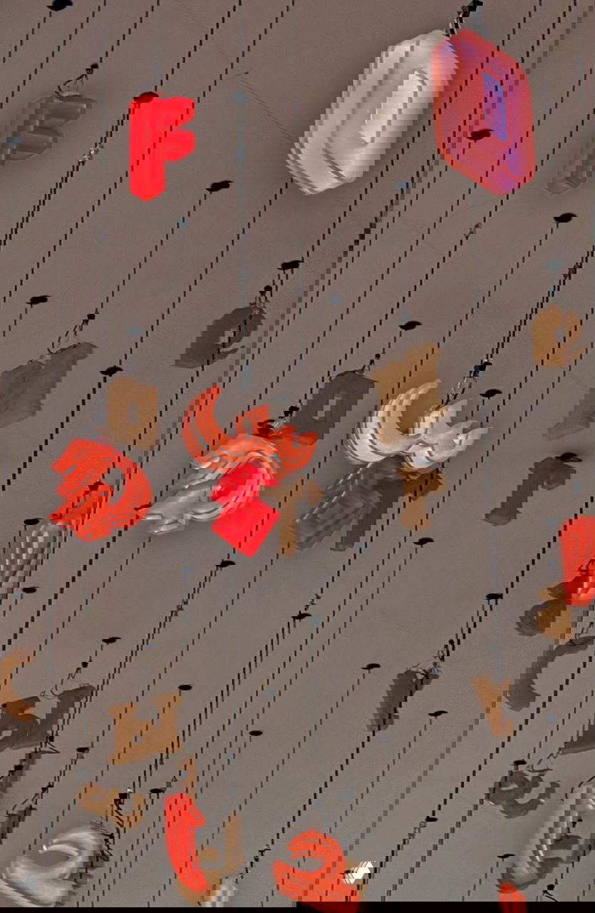 Крупный план подвесных объёмных букв и светящихся элементов инсталляции, свисающих с потолка на длинных тросах над холлом.