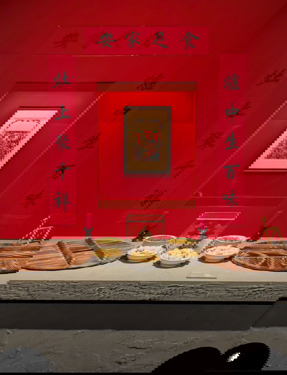 Экспозиция с подношениями и красными декоративными свитками на стене: музейная витрина, где показаны ритуальные блюда и предметы домашнего алтаря.