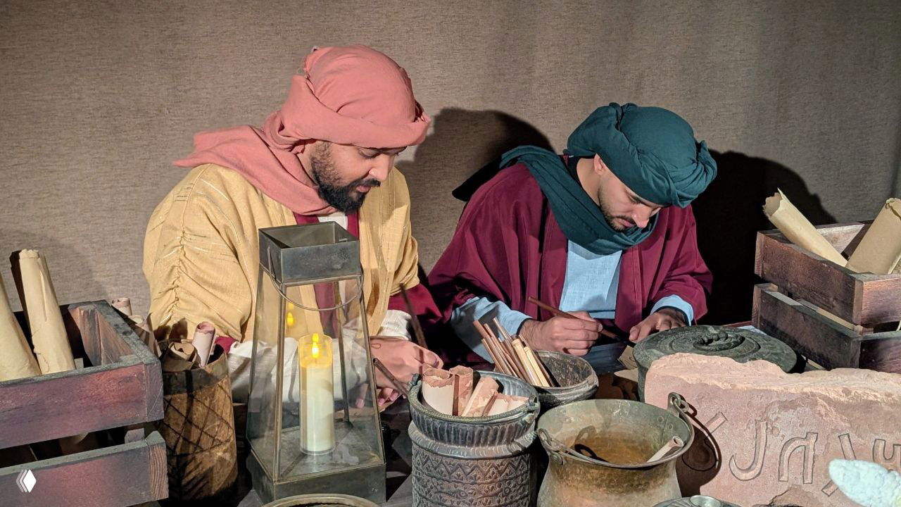 Реконструкция ремесленных сцен: два артиста в традиционной одежде за столом с глиняными сосудами, свитками и лампой в декорации шоу внутри Хегры.