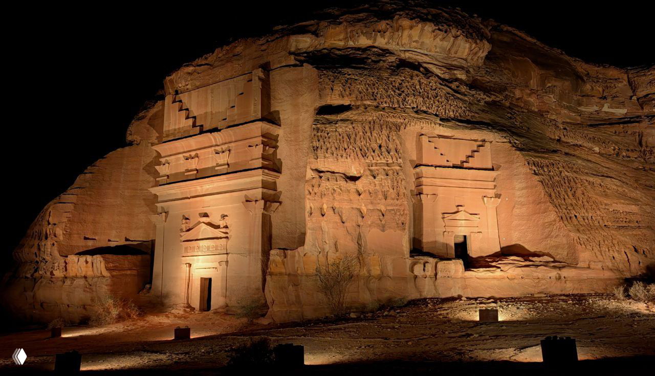 Фасад монументальной гробницы, высеченной в скале, подсвеченный прожекторами на тёмном фоне неба, видно декор и архитектурные детали набатейского стиля.