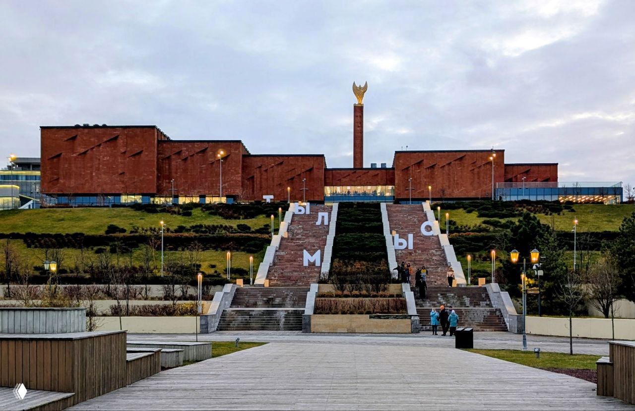 Дневной вид на библиотеку со ступенями и спуском по деревянной набережной; здание на холме видно в контексте окружающего ландшафта.