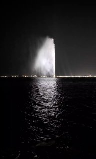 Дальний ночной кадр: фонтан короля Фахда в Джидде как высокая сверкающая колонна воды, на фоне темной гавани и мерцающих огней.