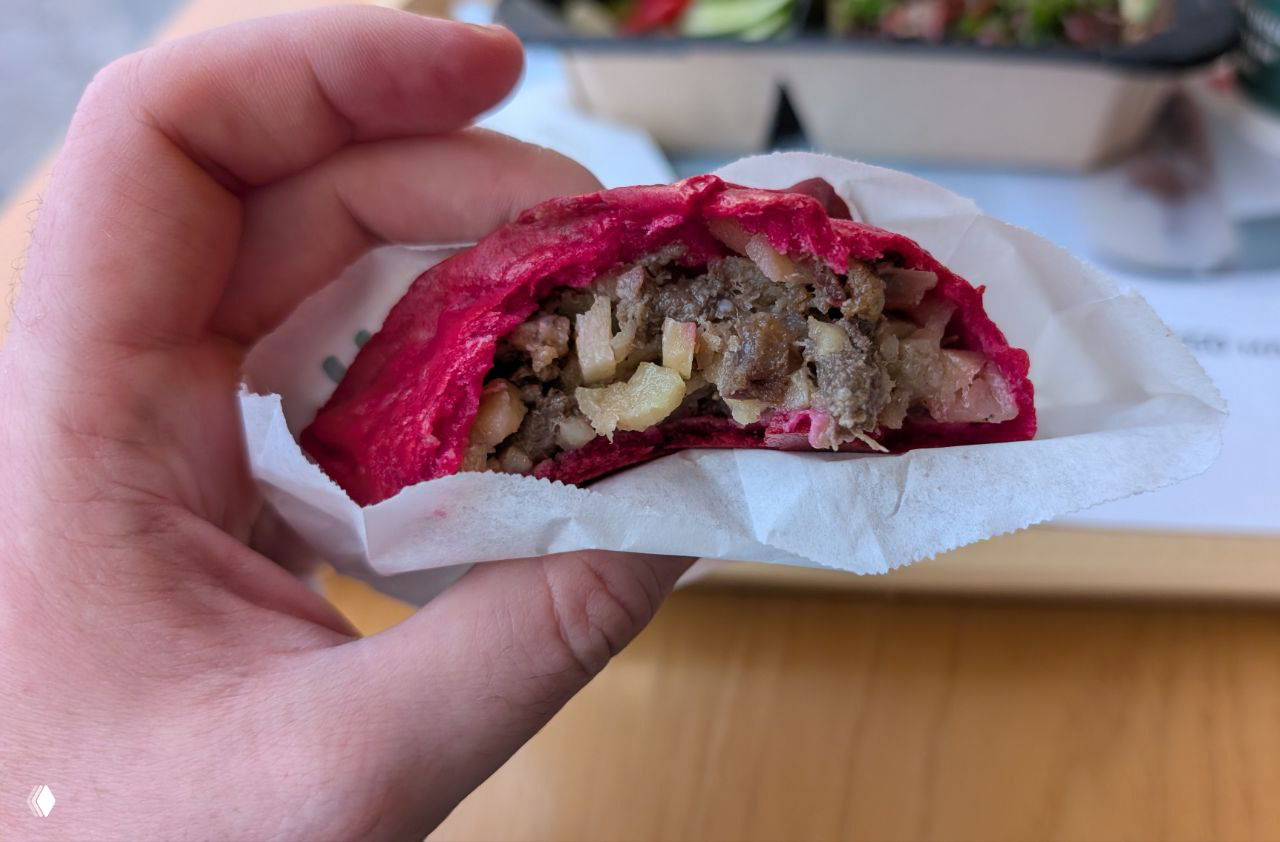 Рука держит розовый пирожок с надкушенным «крашеным» эчпочмаком с кониной: видна мясная и картофельная начинка, бумажная обёртка и интерьер точки на заднем плане.