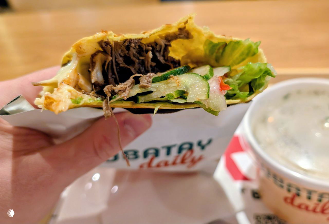 Крупный план начатой лепёшки/лаваша с мясной начинкой, свежими овощами и зеленью, держится в бумажной упаковке с частично видимым логотипом заведения.