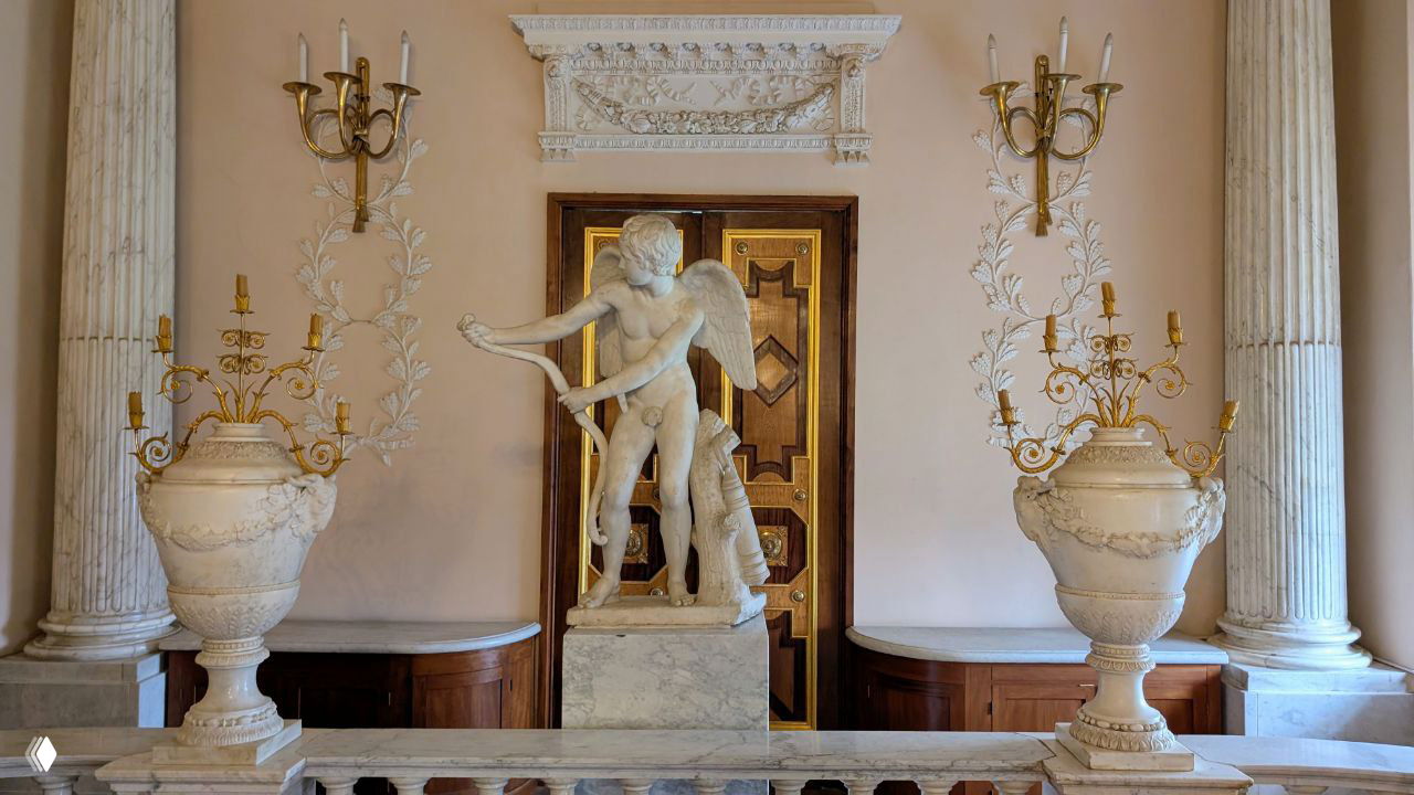 Мраморная декоративная композиция с фигурой амура и парными ваза-урнами на фоне колонн и рельефной отделки интерьера дворца.