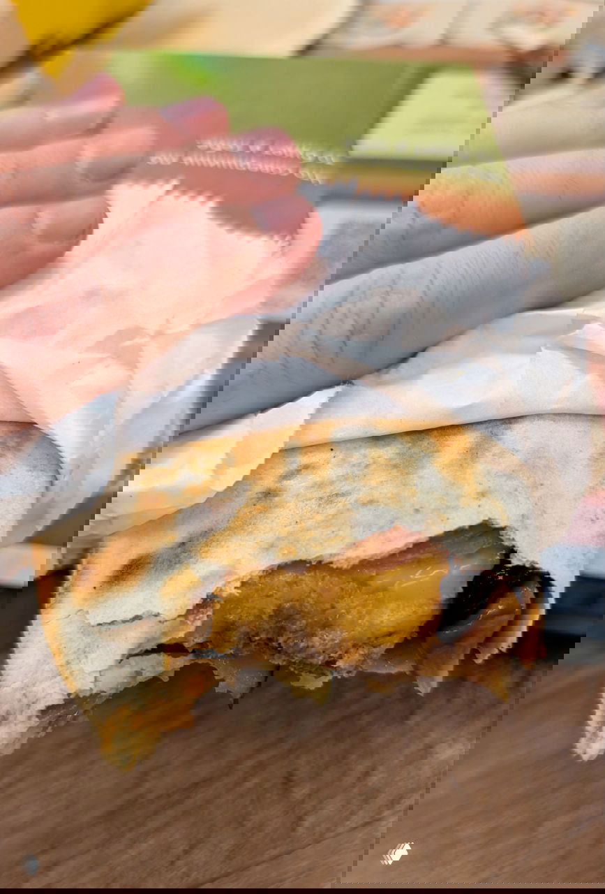 Крупный план надкусанного ролла/лаваша с тянущимся сыром и мясной начинкой, бумажная обёртка в руке автора, видна консистенция и соус внутри.