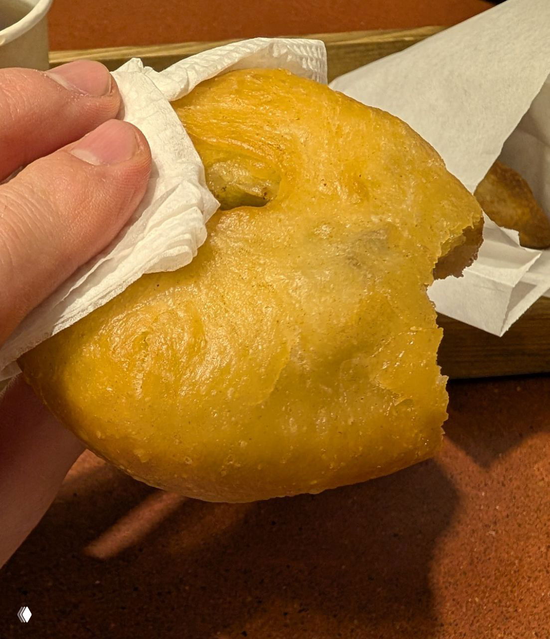 Крупный план жареного пирожка — горячий перемяч/беляш: золотистая хрустящая корочка, откушенный край и бумажная салфетка в руках автора, видна текстура теста.