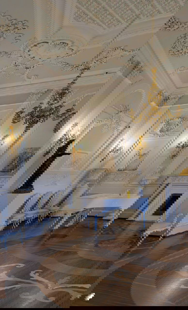 Светлая гостиная с белым роялем, изящной лепниной на стенах и сложным орнаментом паркетного пола в интерьере дворца.