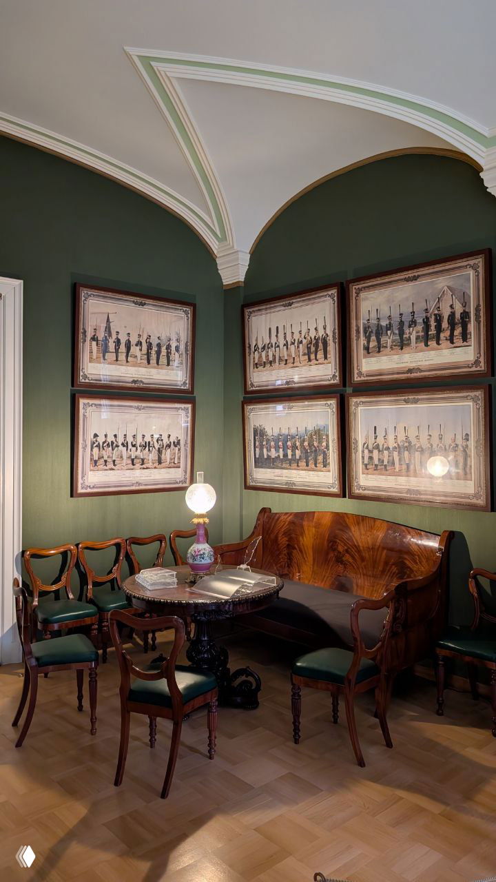 Уютный уголок с кожаной софой, резным деревянным столиком и витиеватым потолком, на стенах несколько рам с военными сценами — интерьерный фрагмент Гатчинского дворца.