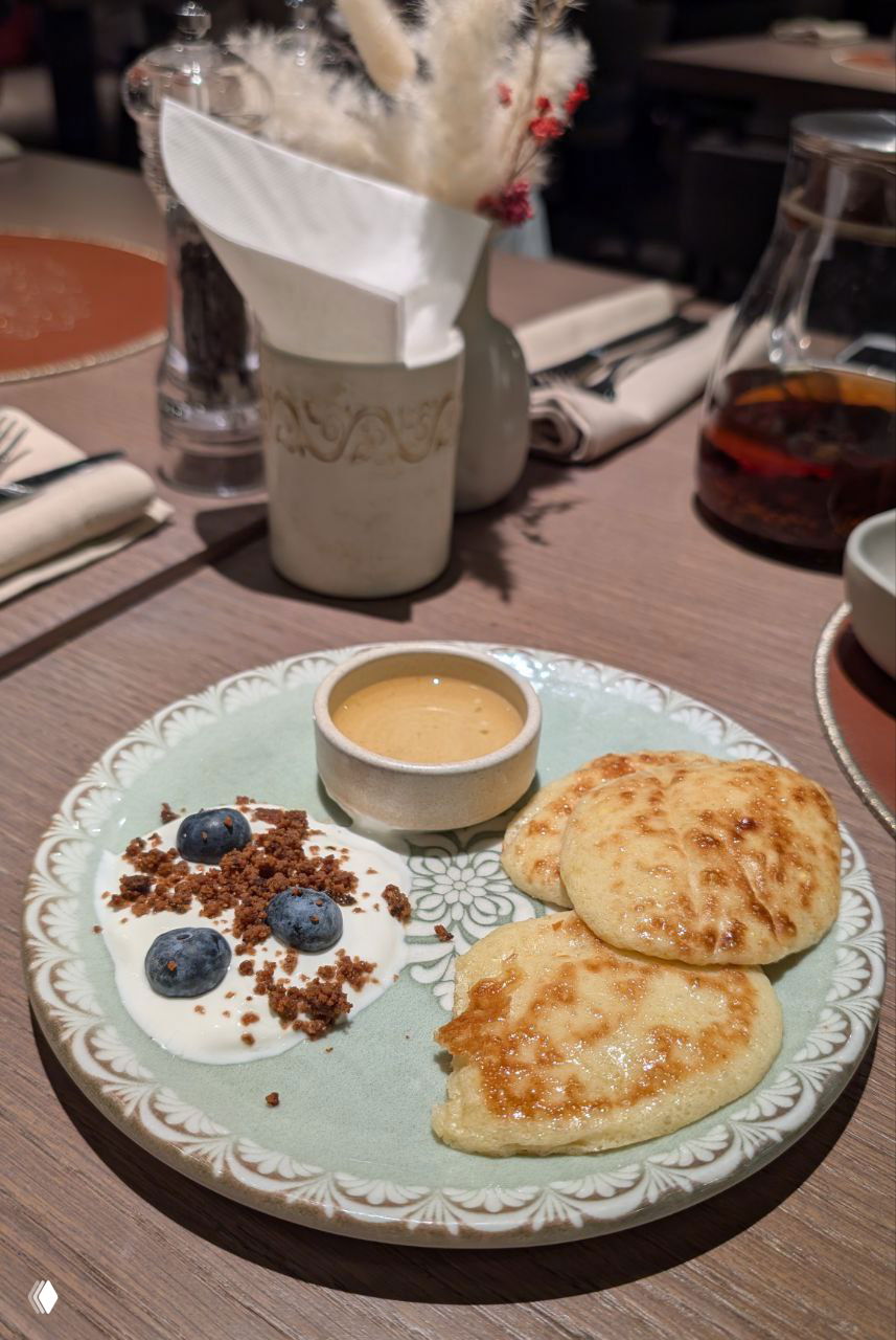 Тарелка с коймаками: три пышные оладьи, рядом ягодный гарнир и соус в небольшой чашке, подача на декоративной керамической тарелке.