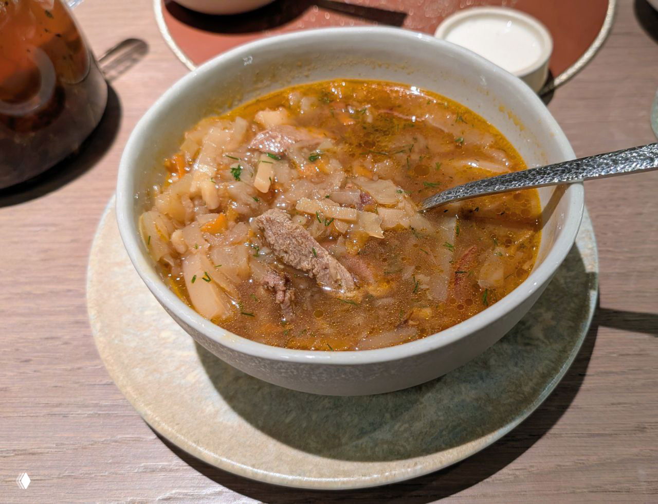 Глубокая миска с наваристым супом: бульон с крупинками, кусочками мяса и овощами, сервировка на столе рядом с чашкой чая.