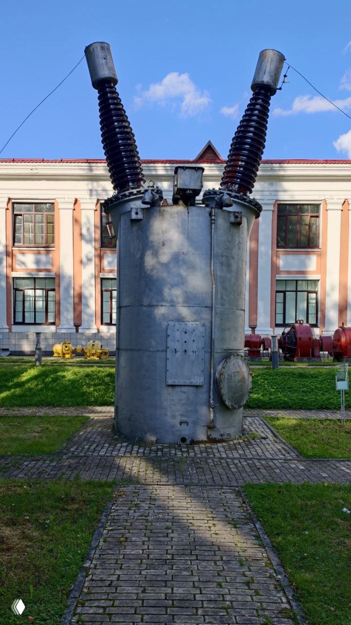 Крупный кадр старого трансформатора или электротехнического агрегата на территории музея на фоне музейного корпуса и зелёной лужайки.