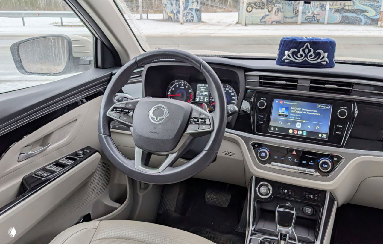 Интерьер и центральная консоль KGM Korando: рулевое колесо, приборная панель и большой экран с Android Auto, тюбетейка на торпеде в кадре.