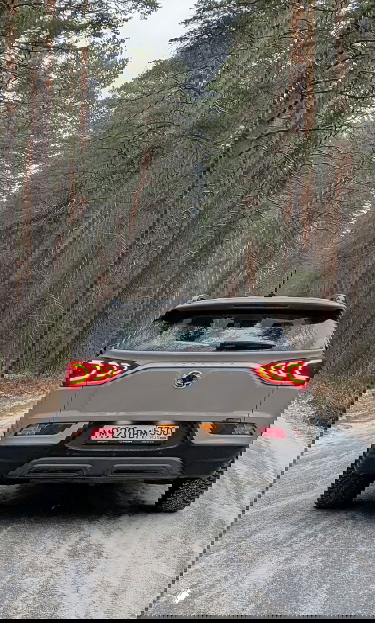 Задний вид KGM Korando на снежной лесной дороге, включённые задние фонари, светлый кузов и следы снега на стекле и бампере, зимний пейзаж.