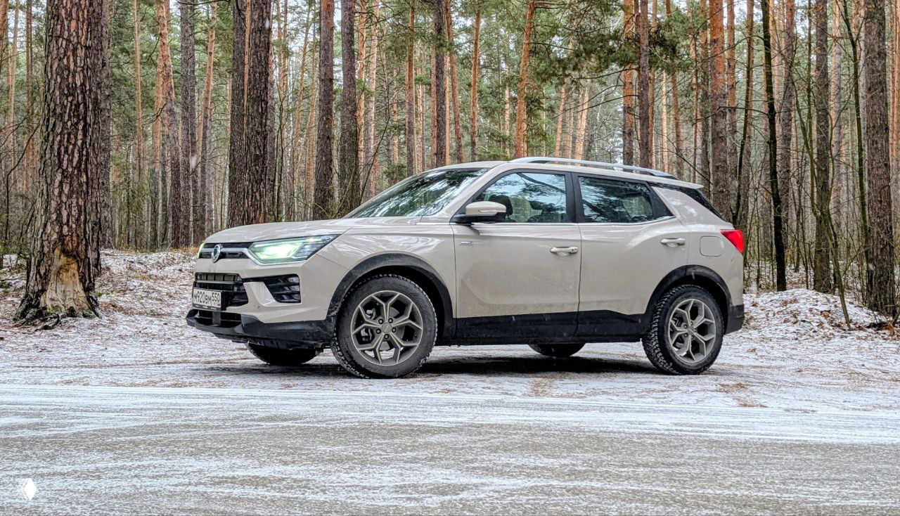 Боковой профиль KGM Korando на лесной поляне в лёгком снегу, автомобиль на зимних шинах, видны клиренс и дизайн колёс, окружение сосен.