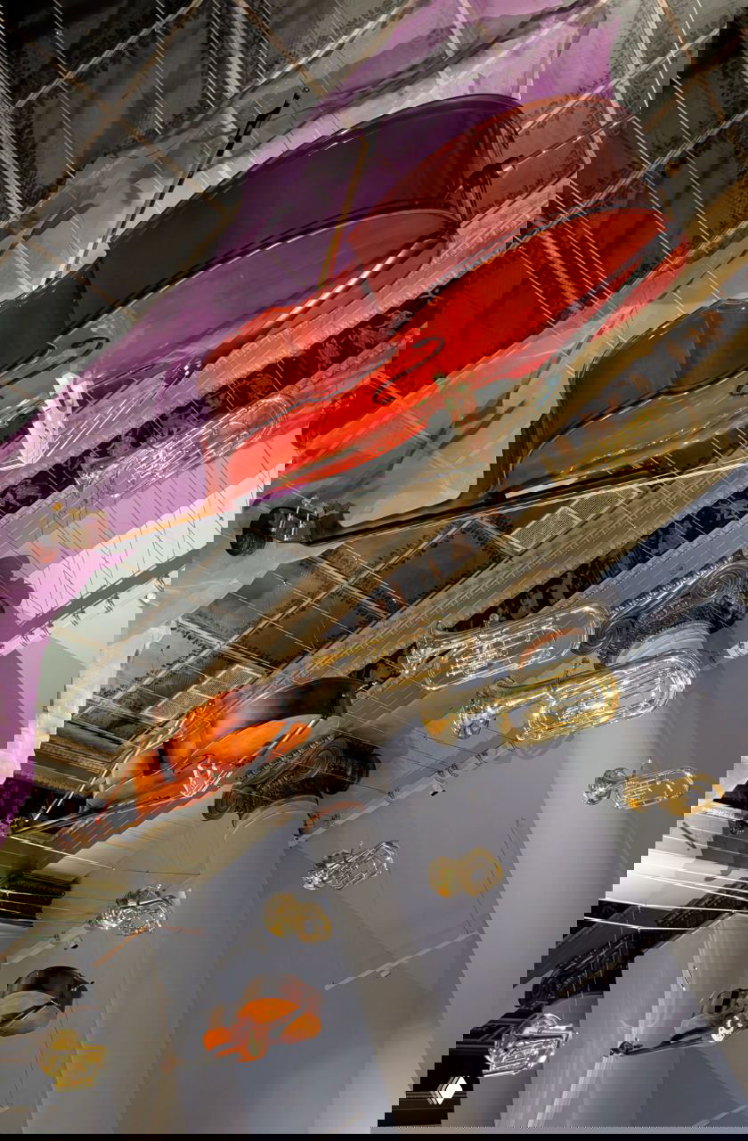 Крупный план подвешенной виолончели в инсталляции: тёплое дерево, отражения света и фиолетовая ткань на фоне музейных колонн.