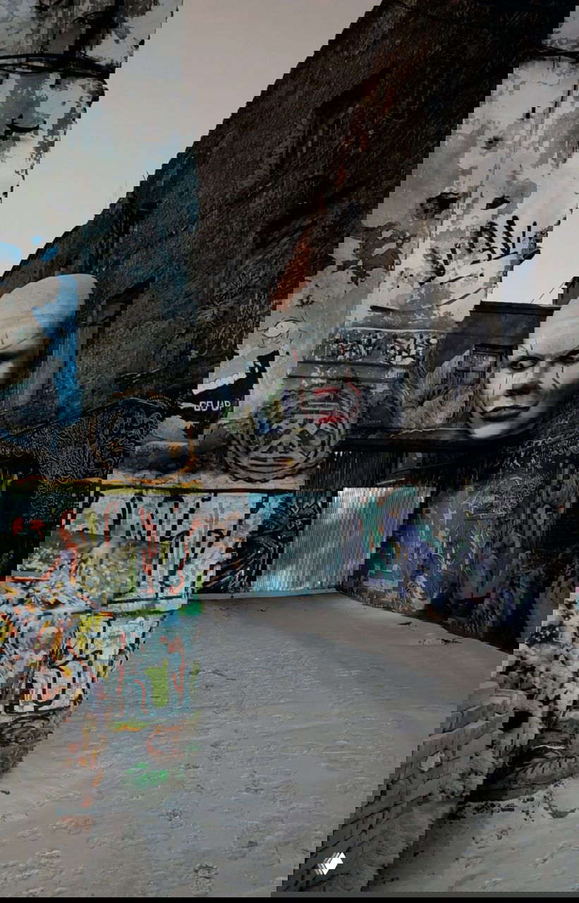 Узкий проход между стенами фабрики с большой зловещей маской клоуна, граффити по бокам и заснеженной землей — ночная атмосфера.