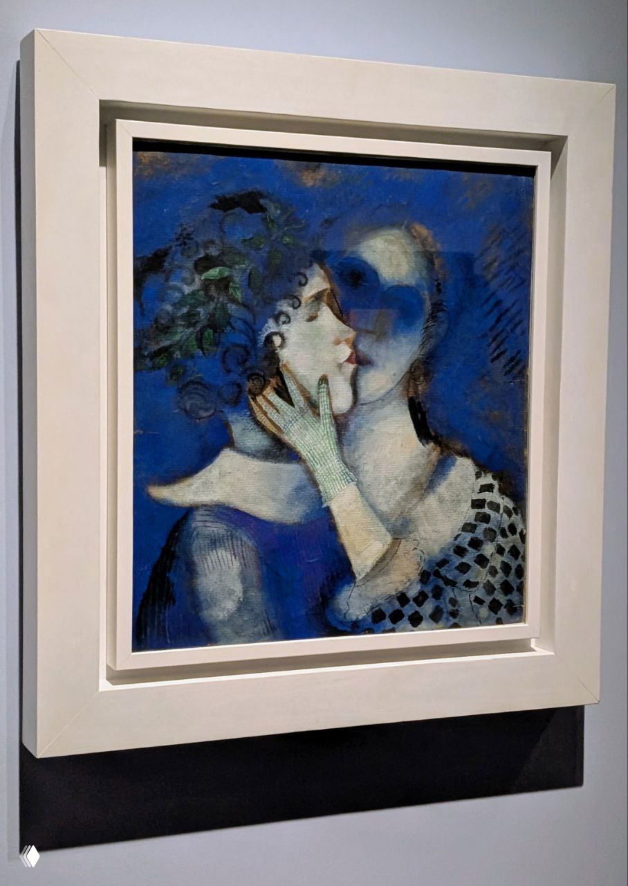 Сине-голубая работа Шагала: поцелуй двух фигур в нежной палитре, видны мазки и текстуры краски в музейной раме.