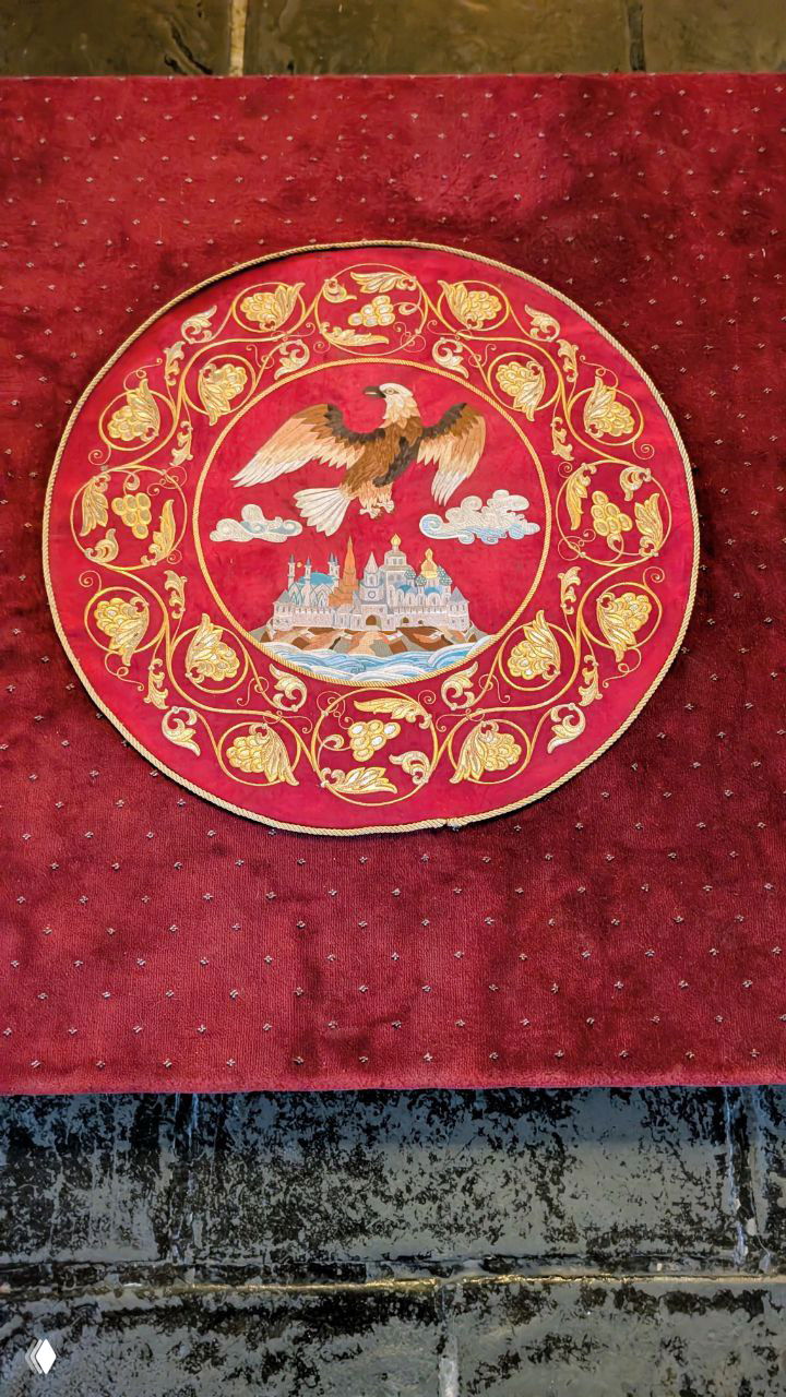 Красный круглый декоративный элемент с золотым орлом и миниатюрным видом кремля в центре — деталь интерьера или церемониального покрытия.