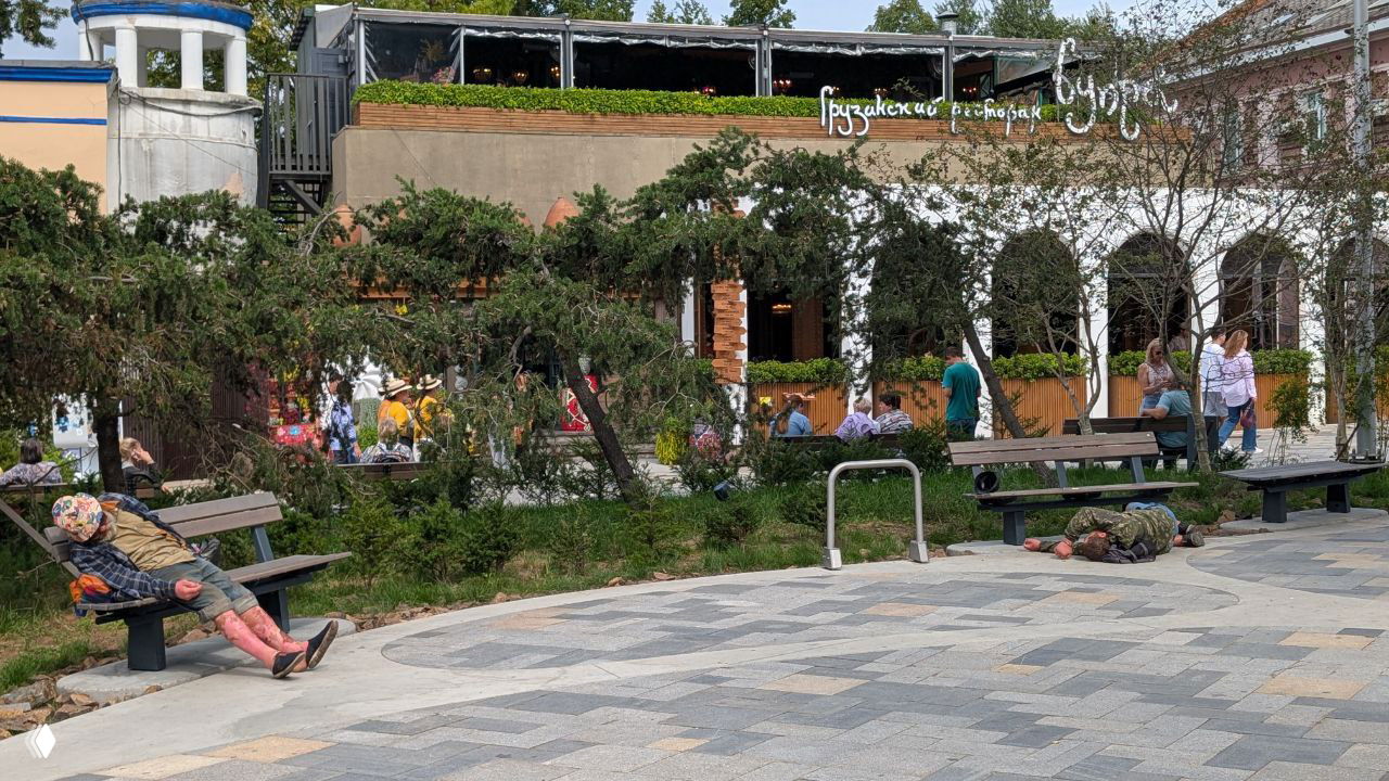 Площадь с лавочками и двумя расположившимися на земле людьми, прохожие и зелёные насаждения на фоне кафе и городской сцены