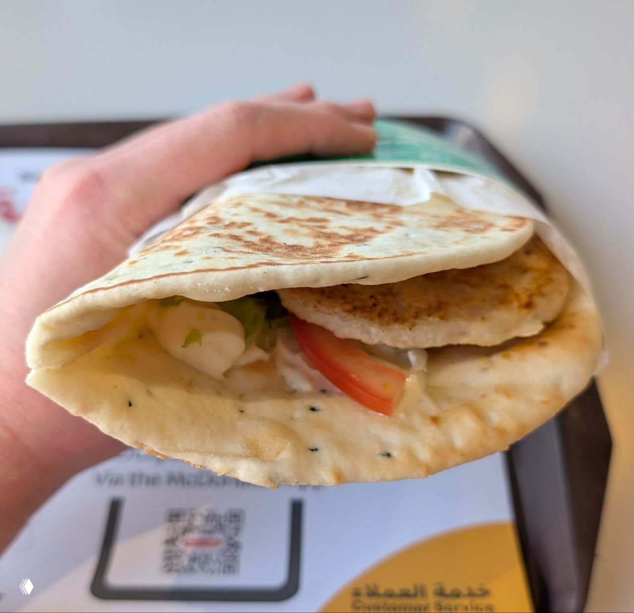 Рука держит большую шаурму в лепёшке из сети Shawarmer: видна начинка, соус и текстура лаваша — уличная еда по местным ценам.