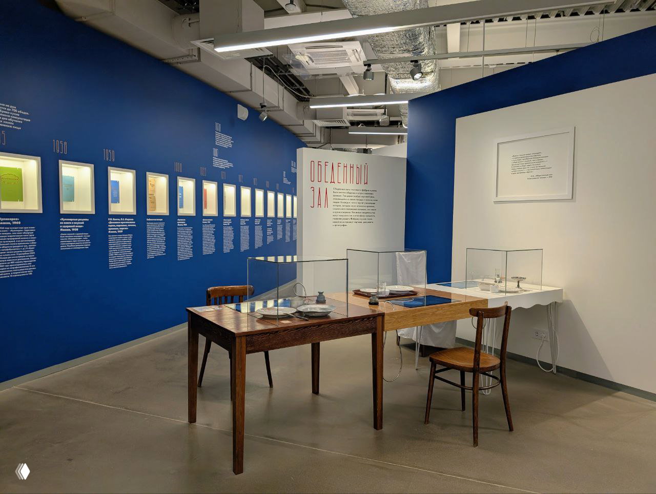 Зал с синими стенами: стол, стулья и витрины с объектами и текстовыми подписями — музейная подача экспозиции Фабрики‑кухни.