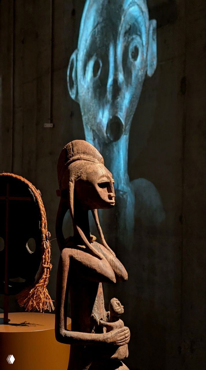 Деревянная скульптура матери с младенцем на руках на фоне крупной проекции лица — музейная инсталляция в зале экспозиции Зиларт.