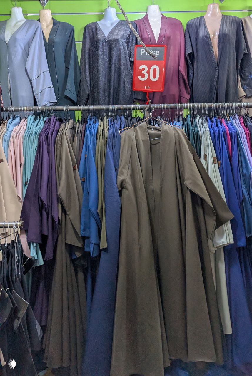 Ряд абай и других женских платьев на вешалках в магазине, красная табличка со скидочной ценой 30 SR и ассортимент тканей и цветов.