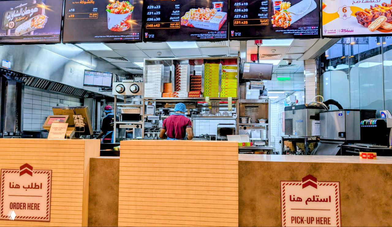 Внутренний вид стойки выдачи в Shawarmer: кухонная зона, экранное меню и сотрудник за прилавком, знак 'PICK-UP HERE'.