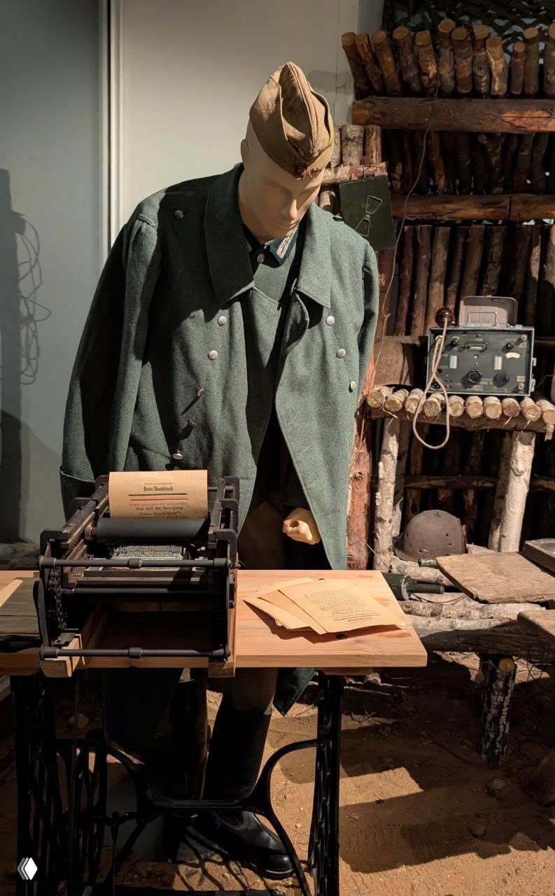 Музейный манекен в военной форме у набора печатного оборудования и полевых принадлежностей — экспонат, иллюстрирующий быт и работу лагерной школы.