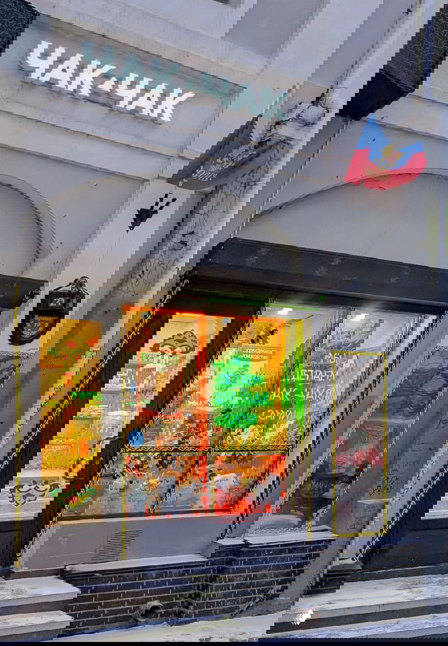 Небольшой уютный вход в магазинчик с неоновой вывеской «ЧАК-ЧАК» и стеклянной витриной, где продают традиционный татарский сладкий чак‑чак.