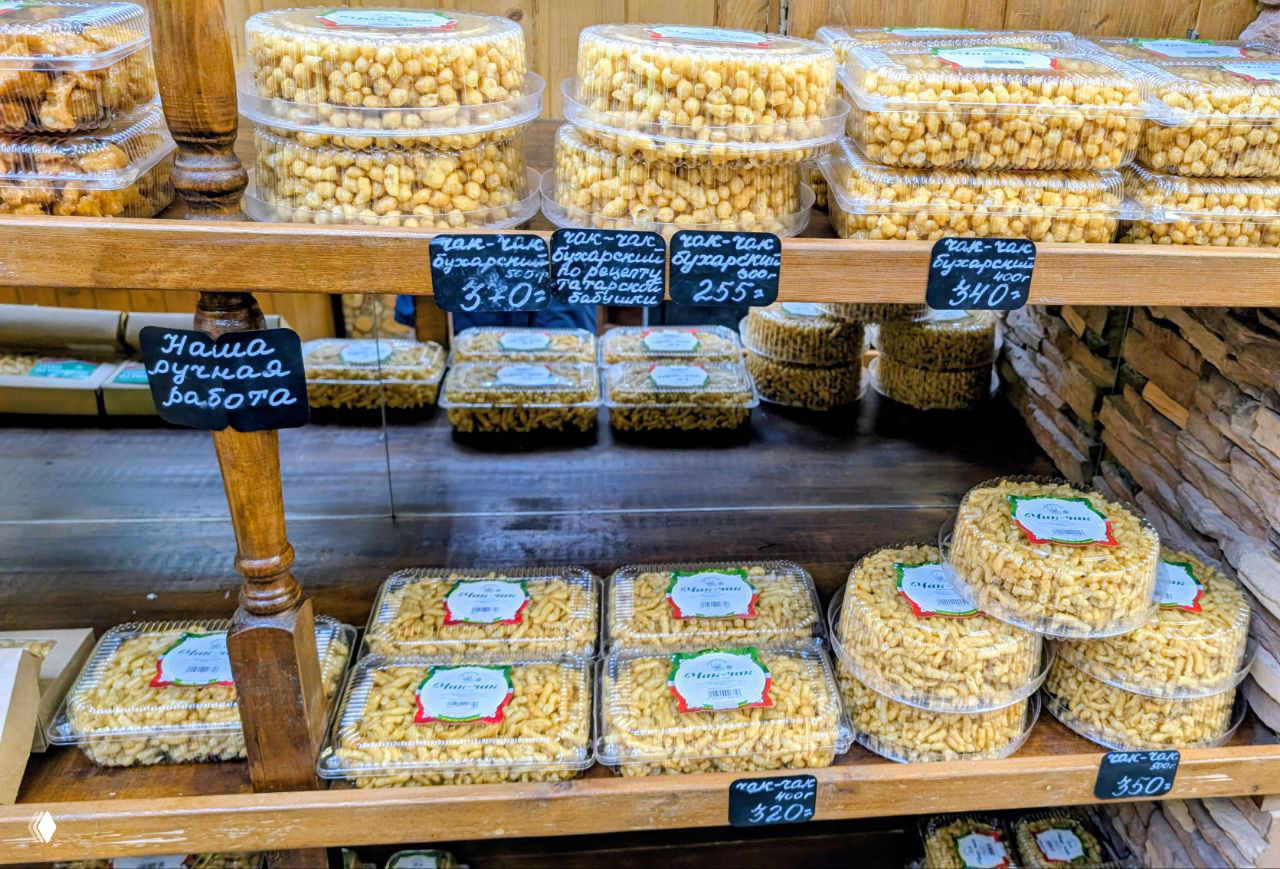 Полки с упакованным чак‑чаком в пластиковых контейнерах разного веса и ценниковыми табличками, снимок внутри магазинчика на Баумана.