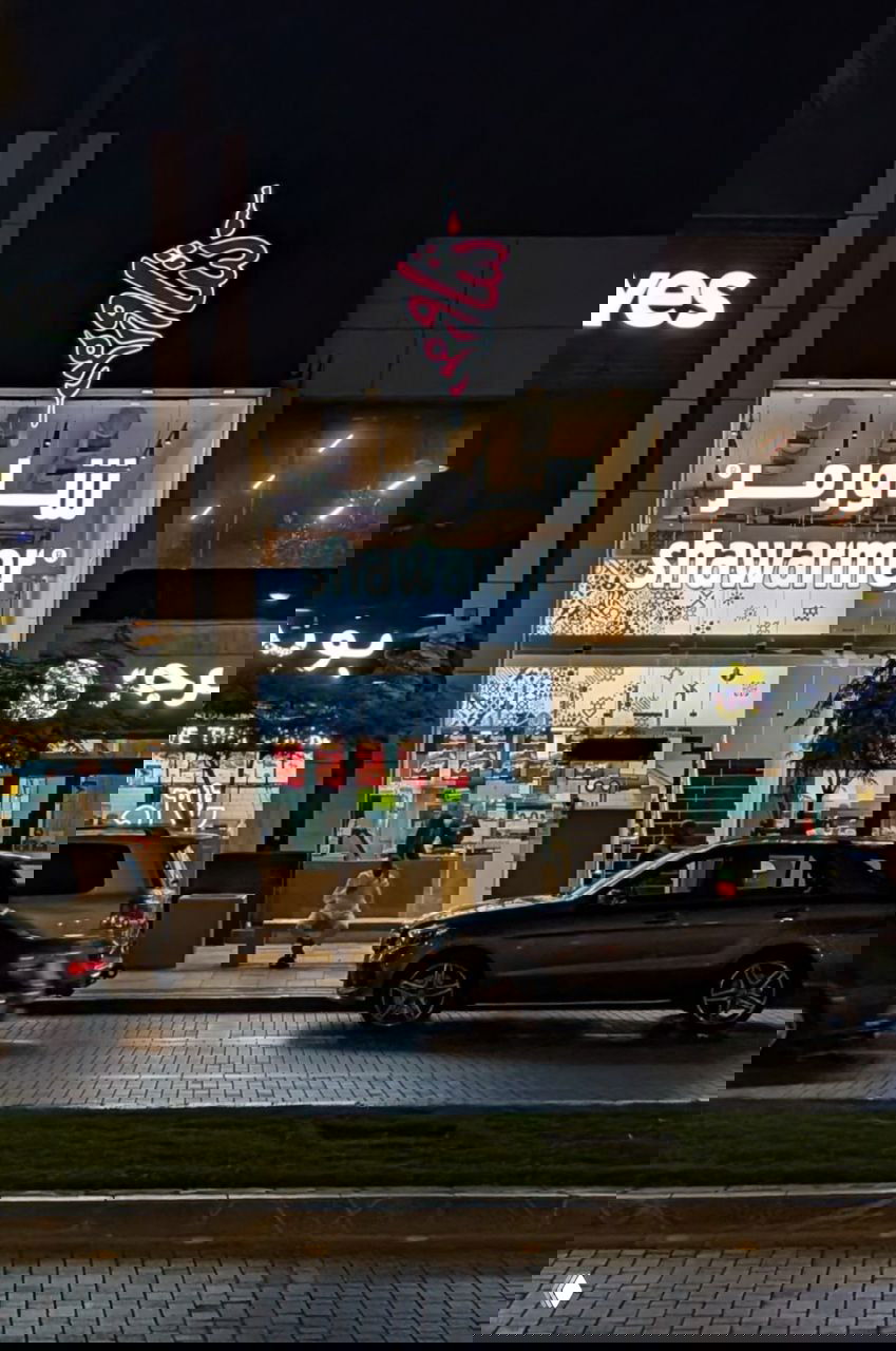 Фасад Shawarmer с двухэтажным остеклением и яркой подсветкой, вид с противоположной стороны улицы вечером.