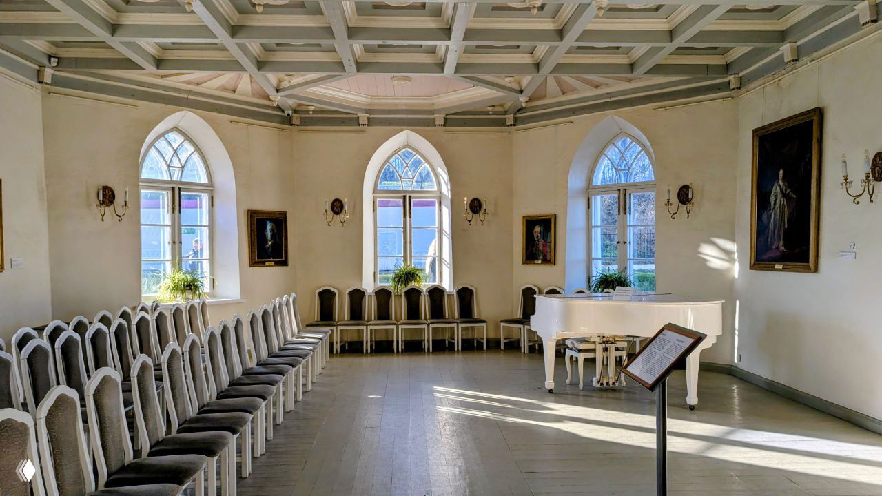 Интерьер светлого зала дворца с белым роялем, рядами стульев по стенам и лучами дневного света, падающими через готические окна.