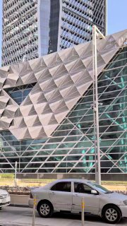 Деталь фасада башни Государственного инвестиционного фонда в Эр-Рияде: металлическая геометрическая облицовка, стекло и припаркованные автомобили у основания.