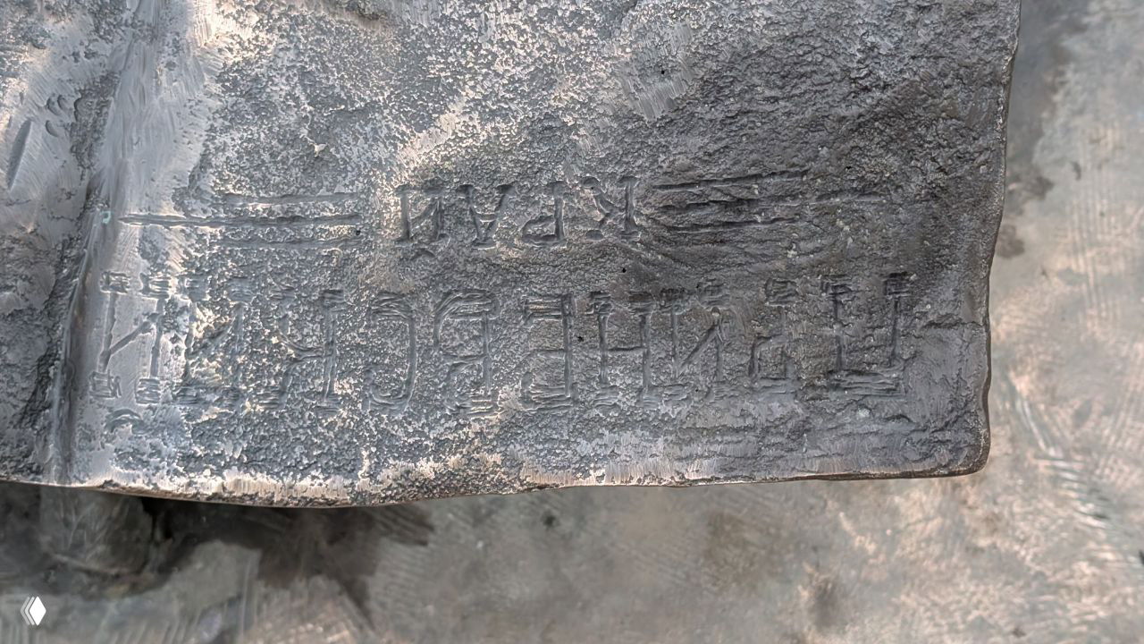 Крупный план выгравированной подписи и текстуры бронзовой скульптуры — видны буквы и следы художественной обработки металла.